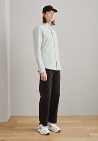Camicia button-up verde chiaro, pantaloni neri a gamba larga, sneaker bianche e cappellino nero; tessuto liscio, design elegante e colori a contrasto.