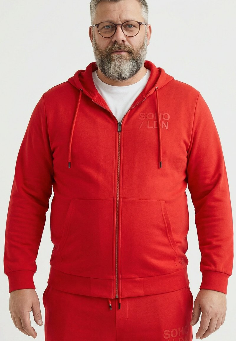 Uomo di mezza età con occhiali e barba che indossa una felpa con zip rossa e pantaloni rossi abbinati su uno sfondo bianco semplice.