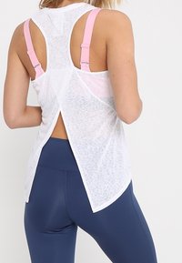 Débardeur de sport blanc avec bretelles roses, doté d'un design dos ouvert et d'une texture subtile. Associé à des leggings bleu marine.