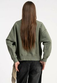 Osoba z długimi brązowymi włosami, ubraną w za dużą zieloną bluzę i czarne dżinsy, stojąca z rękami za plecami na białym tle.