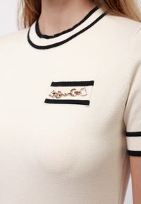 Top en tricot crème avec des bordures noires au col et aux manches, présentant une petite poche sur la poitrine avec un détail en chaîne dorée.