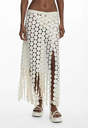 Žena v bielej sukni vyrobenej z prepojených kruhových diskov s vysokými rozparkami, doplnená bielymi platformovými sandálmi.