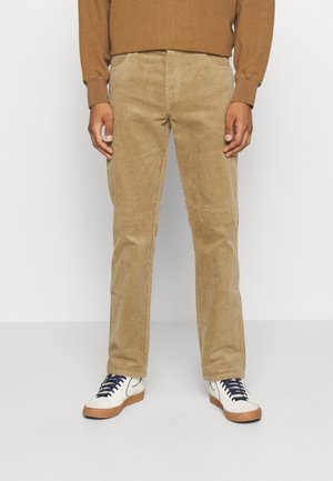 Pantalon en velours côtelé beige avec des côtes verticales, coupe droite, et poches avant, associé à des baskets basses blanches avec des lacets bleu marine.