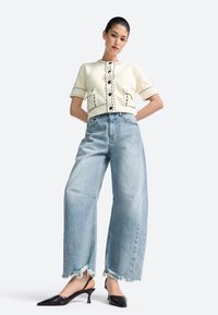 Mujer con cárdigan corto de punto color crema y botones negros, vaqueros anchos de lavado claro con bajos deshilachados y tacones destalonados negros.