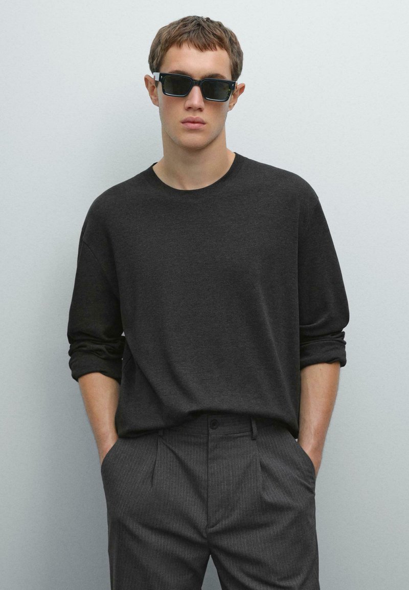 Donkergrijze lange mouwen t-shirt gecombineerd met gestreepte broek. Het shirt heeft een ronde hals en een ontspannen pasvorm. Vierkante zwarte zonnebril gedragen.