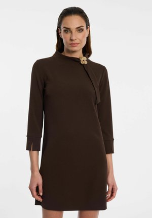 Woman wearing a dark brown long-sleeve dress with a high neckline and decorative gold clasp on one shoulder.