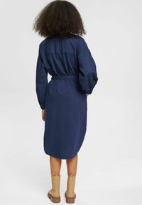 Abito blu navy a maniche lunghe con vita cintrata, orlo a ginocchio e texture sottile. Indossato con stivaletti beige alla caviglia.