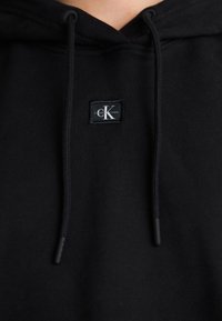 Black hoodie made of soft fabric, featuring a front pocket, drawstrings, and a square logo patch with "Calvin Klein Jeans" in white lettering.