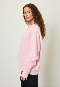 Vaaleanpunainen löysä neulepaita, jossa on pyöreä pääntie, ribatyt hihat ja pehmeä, tekstuuri päällinen. Yhdistetty tummiin farkkuihin.