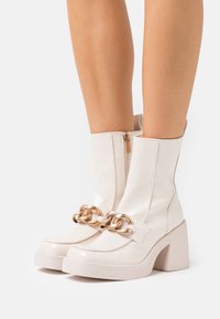 Valkoinen lakattu nahkasaappaat nilkkapituisina, joissa on paksu korko, kultaketjutehoste ja sivuvetoketju suljettavaksi. Tasainen pinta.