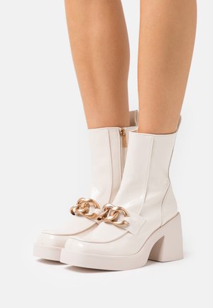 Botas de tobillo de charol blanco con tacón grueso, que presentan un acento de cadena dorada y un cierre con cremallera lateral. Textura suave.