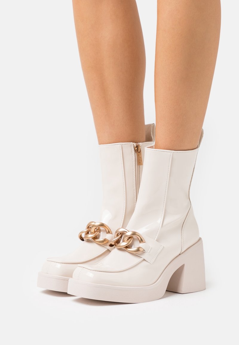 Valkoinen lakattu nahkasaappaat nilkkapituisina, joissa on paksu korko, kultaketjutehoste ja sivuvetoketju suljettavaksi. Tasainen pinta.