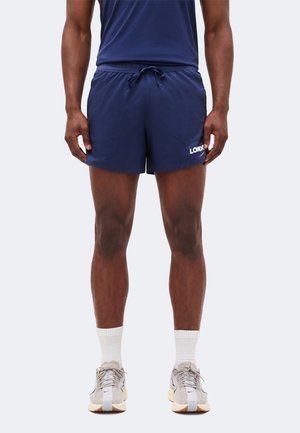 Mężczyzna noszący granatowe sportowe spodenki z napisem "LONDON", białe skarpetki za kostkę i jasnoszare buty do biegania stoi na jednolitym tle.