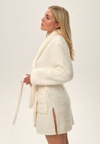 Krótki, kremowy szlafrok wykonany z puszystego materiału, z paskiem, dwiema bocznymi kieszeniami i rozcięciem do wysokości uda. Miękka tekstura i luźny krój.