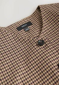 Brown patterned fabric with small diamond shapes in beige and dark colors. Features a black button and a v-neck design. Brand label visible.
