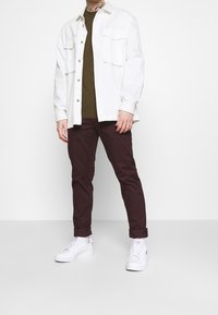 Witte overhemd met knopen en twee borstzakken, olijfgroen getailleerd shirt eronder, bordeauxrode smalle broek en witte sneakers.