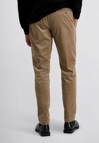 Man in beige corduroy broek, zwarte schoenen en een zwart longsleeve shirt, staand met één hand in zijn zak tegen een effen achtergrond.