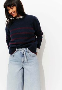 Pull marin à rayures bordeaux, col rond, avec des poignets côtelés. Pantalon en jeans bleu clair à jambes larges avec taille à cordon. Fond blanc.