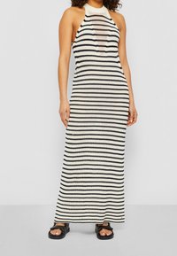 Robe maxi en tricot à rayures crème et noire, présentant un col halter et des fentes sur les côtés, associée à des sandales plates noires.