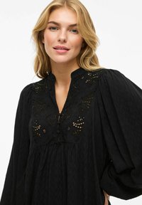 Blusa nera con tessuto testurizzato, collo alto con dettagli in pizzo e maniche a sbuffo. Include accenti di bottoni e una vestibilità sartoriale.