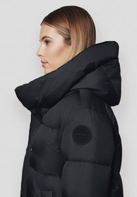 Czarna puchowa kurtka z wysokim kołnierzem i okrągłą łatą z logo na ramieniu. Posiada pikowaną strukturę i dopasowany krój.