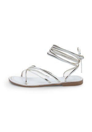 Sandales plates à lanières avec des brides métalliques argentées, une semelle intérieure blanche, un design ouvert et un lacet noué autour de la cheville, reposant sur une semelle marron clair.