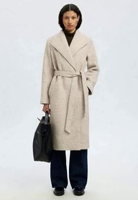 Beige wikkeljas met een strikriem, gekartelde kraag en lange mouwen, gecombineerd met brede, donkere jeans en een zwarte tote tas. Gladde textuur.