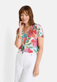 Top de manga corta y cuello en V con un colorido patrón de hojas tropicales en rosa, turquesa y verde sobre un fondo claro. Combinado con pantalones blancos.