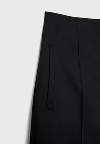 Gros plan d’un pantalon noir montrant la ceinture et la poche latérale verticale à couture soignée sur fond clair.