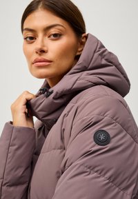 Giacca imbottita in viola tenue con colletto alto e cappuccio regolabile. Presenta una patch logo nera sulla manica e chiusura con zip.