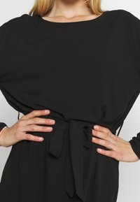 Černé šaty s volným střihem, dlouhými rukávy a zavázaným pasem. Tkanina se zdá být měkká s hladkým povrchem. Jednoduchý, minimalistický design.