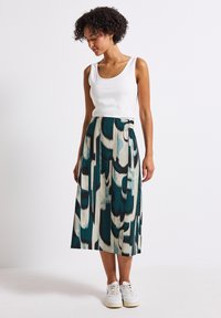 Ein langer, gemusterter Rock in Teal, Schwarz und Creme mit einem strukturierten Stoff. Kombiniert mit einem weißen Tanktop und weißen Sneakers.