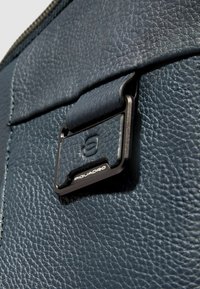 Marineblauw leer met een gestructureerde afwerking. Voorzien van een metalen gesp met het "Piquadro"-logo, wat de hoogwaardige vakmanschap benadrukt.