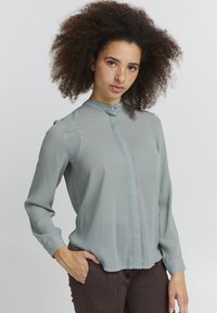 Blusa leggera verde chiaro, trasparente, con colletto alto, chiusura frontale con bottoni e maniche lunghe. Vestibilità leggermente ampia, texture liscia e design semplice.