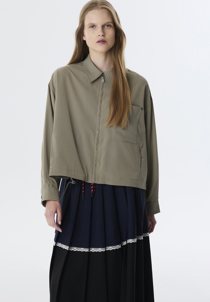 Chaqueta verde oliva con cremallera y cuello tipo camisón, combinada con una falda de navi y negro con capas y pliegues que presenta detalles de encaje.
