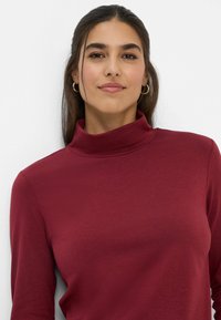 Burgunderfarbener Rollkragenpullover aus weichem Material, mit langen Ärmeln und figurbetontem Schnitt, mit glatter Textur und minimalen Nähten.