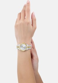 Silberne und goldfarbene Uhr mit einem runden Ziffernblatt, einer strukturierten Lünette und einem gemusterten Metallarmband. Tragen an einem hellhäutigen Handgelenk.