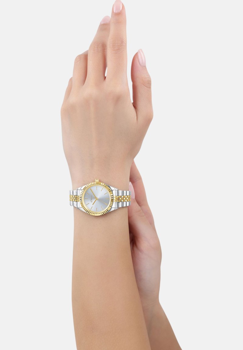 Silberne und goldfarbene Uhr mit einem runden Ziffernblatt, einer strukturierten Lünette und einem gemusterten Metallarmband. Tragen an einem hellhäutigen Handgelenk.