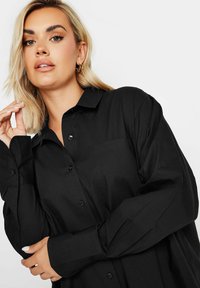 Camisa negra de botones con un cuello estructurado, mangas largas y un detalle de bolsillo. Tela suave con un corte relajado y botones mínimos.