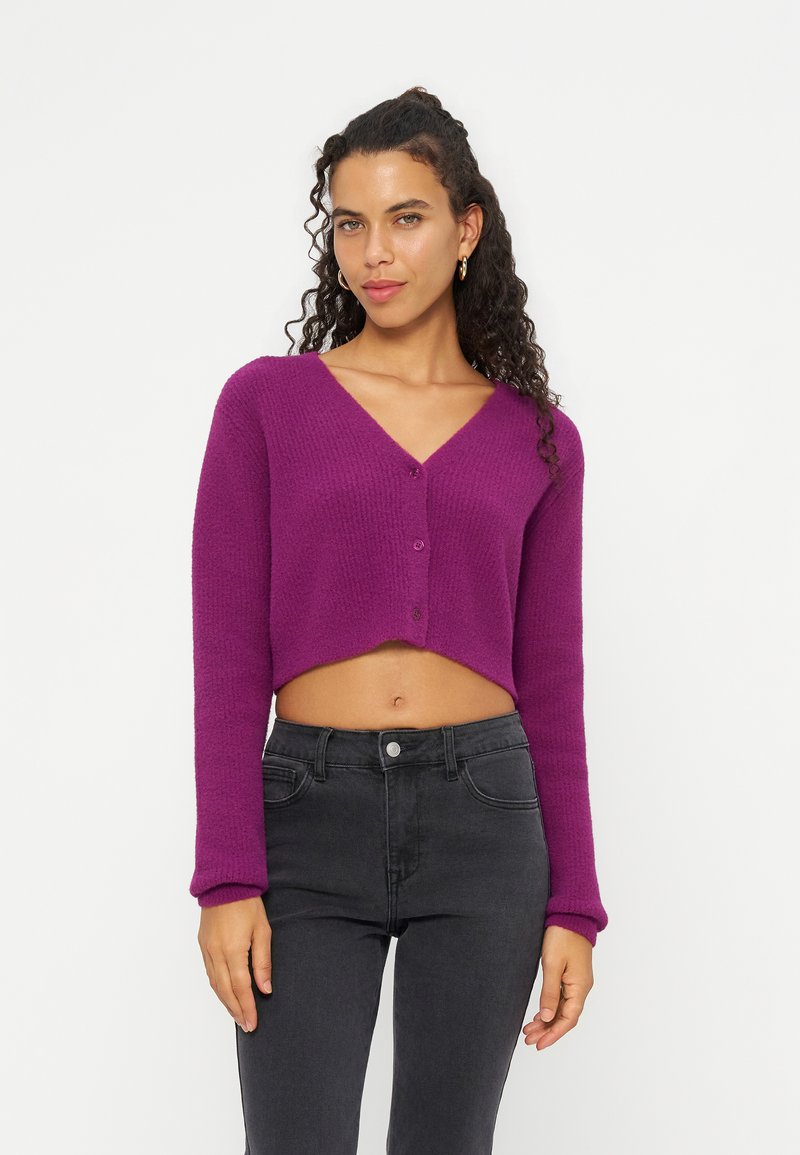 Femme aux cheveux bouclés portant un cardigan violet court et un jean noir taille haute, debout devant un fond blanc uni.