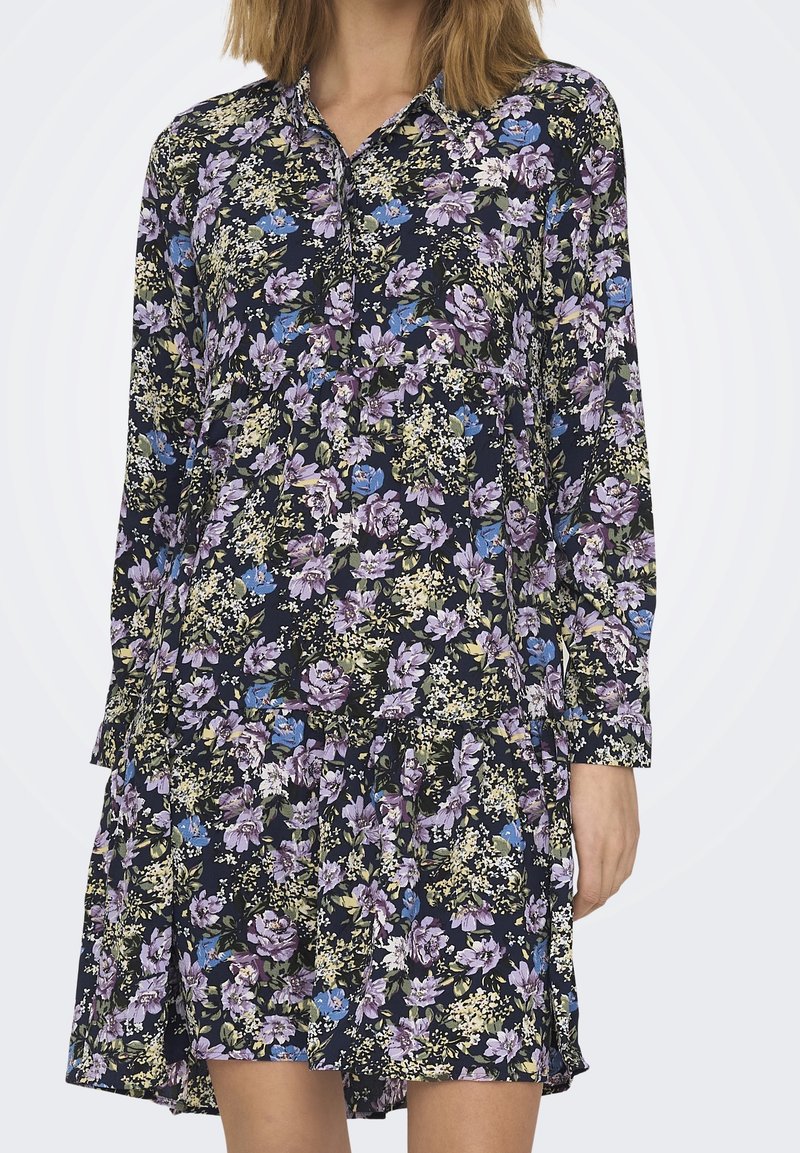 Abito floreale con maniche lunghe, sfondo blu navy, presenta fiori viola, blu e verdi, colletto con bottoni e design a gonna a strati.