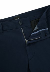 Navyblauwe broek met gedeeltelijk geopend zilveren rits en knoopsluiting, waarbij de binnenste tailleband en zwarte labeltab zichtbaar zijn.