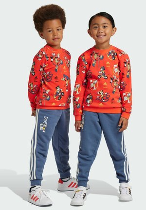 Felpa rossa con stampe di personaggi Disney; pantaloni jogger blu con strisce bianche, dettagli numerici e una texture morbida; entrambi abbinati a sneakers bianche.