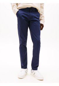 Hombre con pantalones casuales azul marino, zapatillas blancas con rayas negras y un suéter beige, de pie con las manos en los bolsillos.