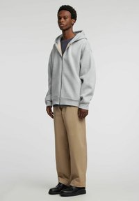 Grå hettejakke med glidelås, stor hette, løs passform og ribbestrikkede mansjetter, kombinert med lys beige vidbukser og svarte sko.