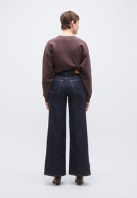 Barna pulóver eresztett vállakkal és széles szárú sötét farmernadrág. Jelentős részletek közé tartozik a látható varrás és a farmer hátulján található címke.