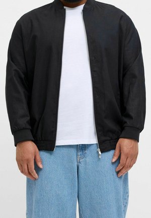 Man wearing a black zip-up jacket over a white shirt with light blue denim jeans, standing against a plain background.