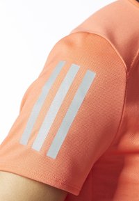 Pomarańczowa koszulka sportowa z fakturowanej tkaniny. Srebrny wzór w trzy paski na lewym ramieniu podkreśla rękaw.