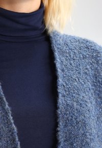 Cárdigan azul texturizado llevado sobre un cuello de tortuga navy. El cárdigan presenta un tejido suelto y un diseño de abertura frontal, enfatizando una apariencia acogedora.