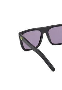 Occhiali da sole neri con lenti viola, design angolare, finitura opaca e un dettaglio floreale verde sul tempio sinistro.